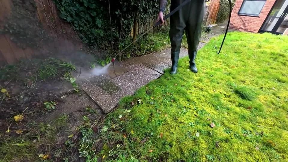 Technician in waders pressure washing a concrete garden path beside a mossy lawn